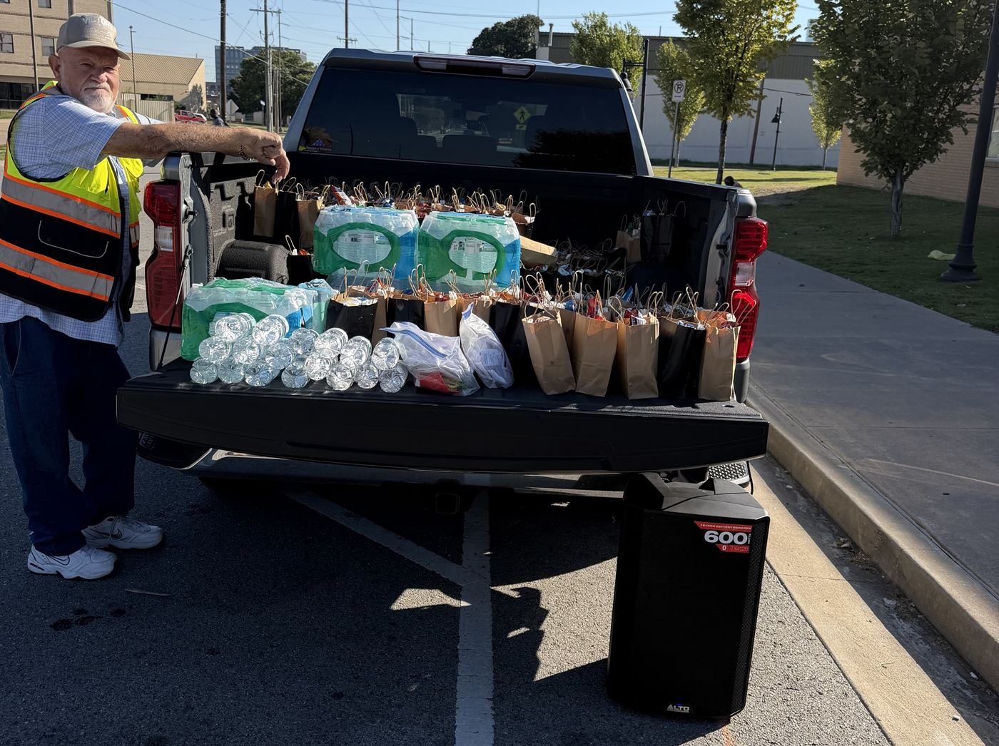 A volunteer in a safety vest beside a tailgate lined with lunch bags, water and a speaker